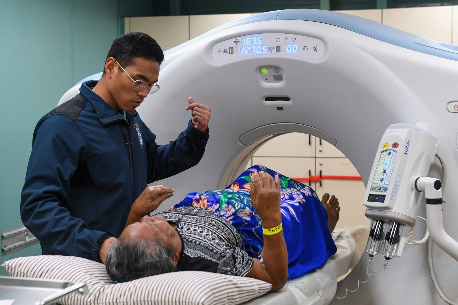 A gray-haired person lies on a CT scanner table wearing their own patterned clothing, with a bright blue floral cloth draped over them. A staff member stands beside the table with one hand raised, explaining something.